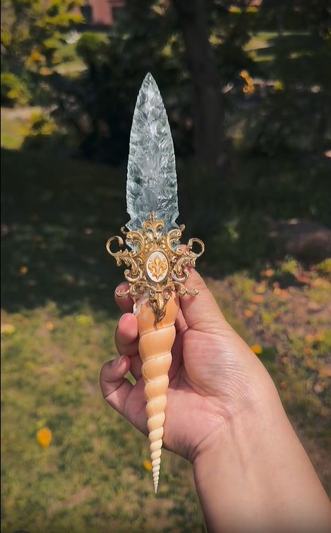 Macro close-up of the dagger's hilt, featuring a genuine ammonite fossil, shimmering abalone shell, and an ornate bronze cap